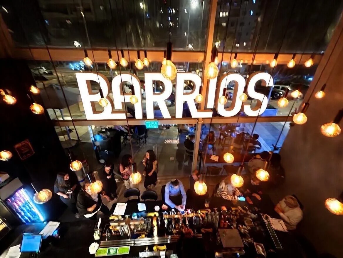 הבר מלמעלה בערב: שלט BARRIOS מואר, נורות תלויות ואנשים ליד הבר